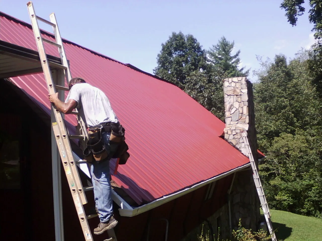 Roofer on a ladder beside a red metal roof and stone chimney — Soffit & Fascia Repair by Roofing Brothers in Mattawa